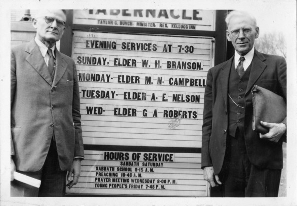 Two unknown men standing in front of the Battle Creek Tabernacle announcement sign