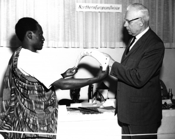 William G. C. Murdoch looks at an art object held by a man from Africa at a missions display