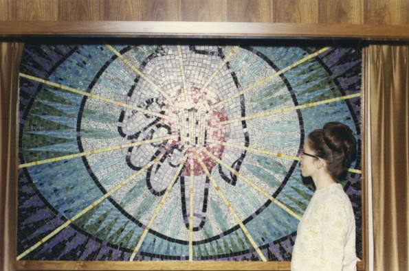 [Ann Steinweg stands beside a mural of her own making. The mural depicts Christ's hand holding the symbol of Andrews University]
