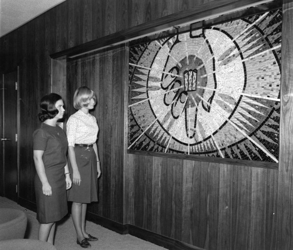 [Two students admire Ann Steinweg's mural of Christ's hand holding the symbol of Andrews University on display at the Andrews University board room]