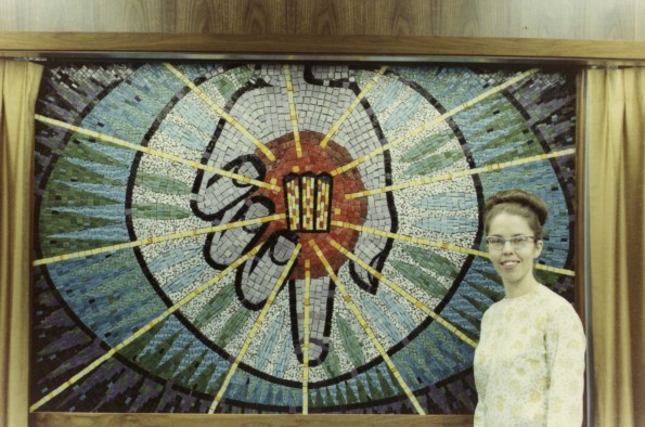 [Ann Steinweg stands beside a mural of her own making. The mural depicts Christ's hand holding the symbol of Andrews University]