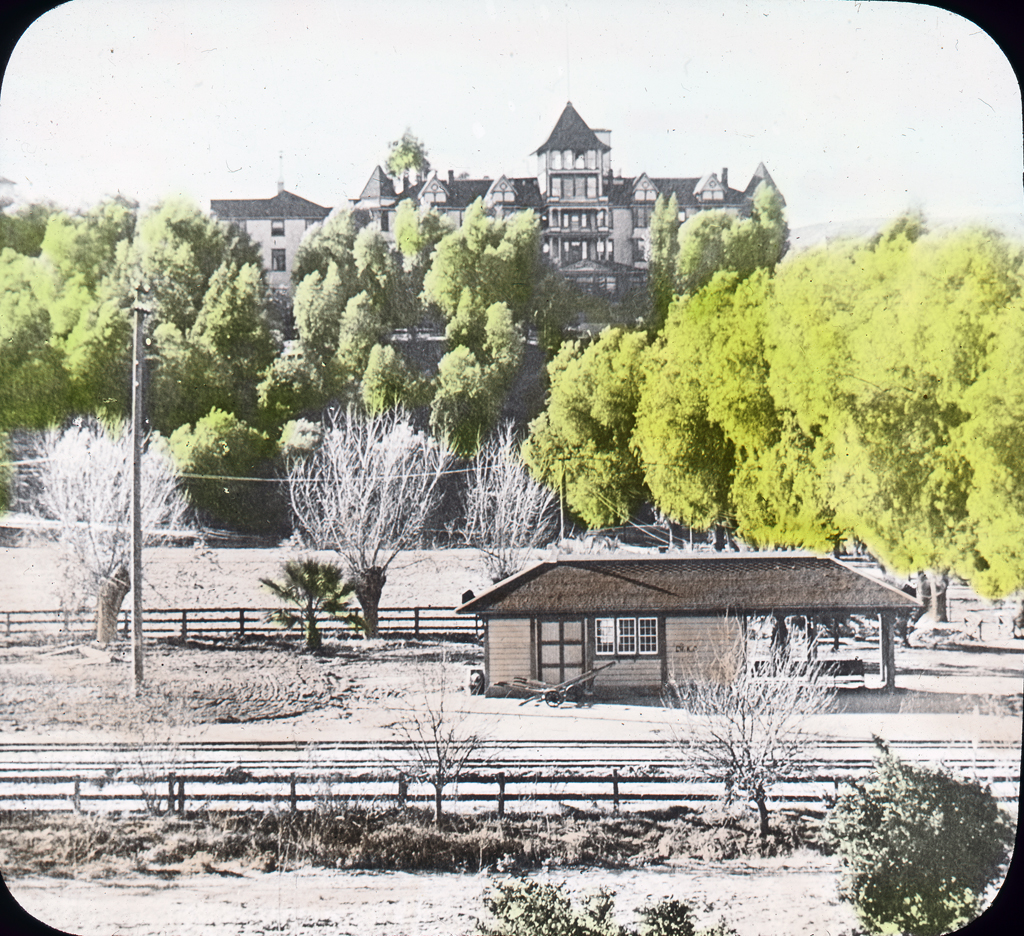 Lantern slide. A washed-out looking view of a clearing with a few shrubs, bushes, and train depot with larger trees in the background. In the distance is the Loma Linda Sanitarium, a long building with a tower.